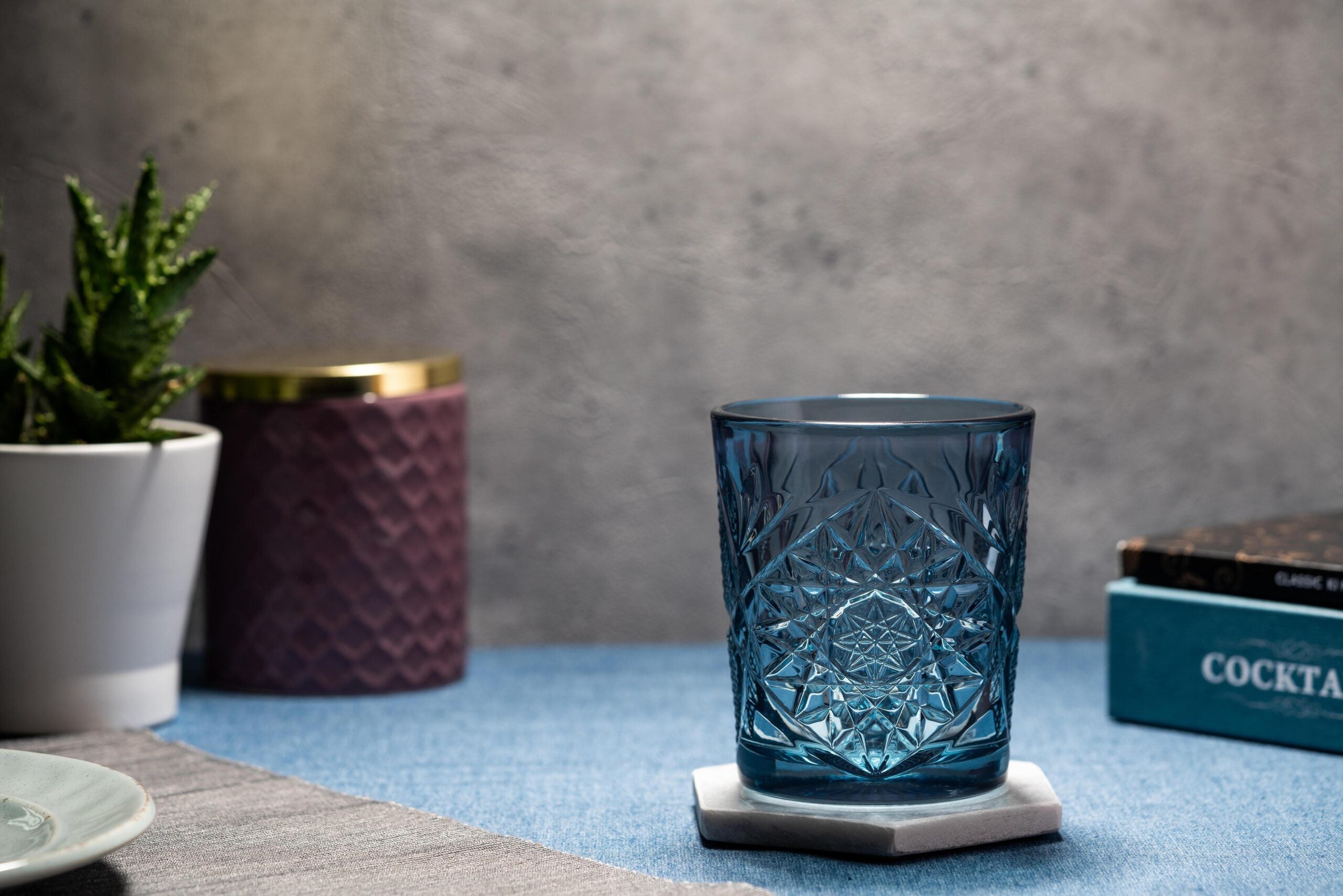 Kuva Hobstar laseista ja vieressä kirja ja viherkasvi. Lasin väri on petroolin sininen.